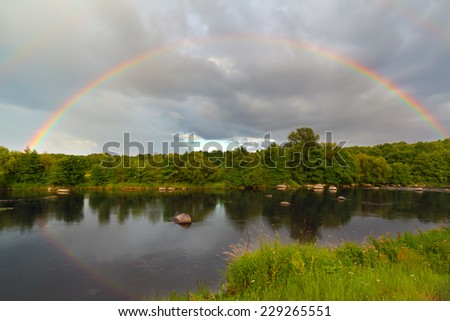 Full rainbow over forest  and river with reflection in water and cloudy sky