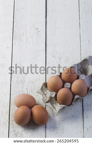 Brown chicken eggs in a gray cardboard tray on textured, painted white boards, lighted studio light.