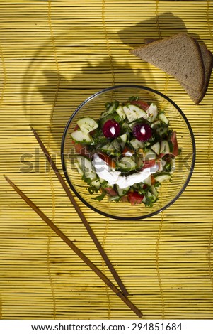 Tomatoes and cucumbers in a glass plate with chopsticks, a piece of rye bread on a yellow bamboo mat on a wooden background, studio lights lit, the view from the top.