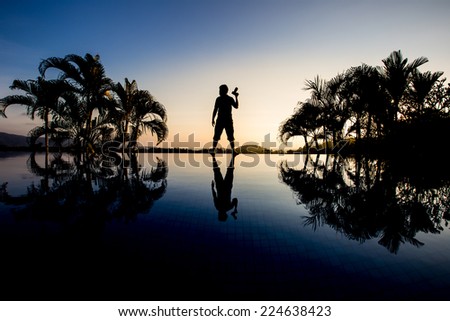 silhouette traveler hold camera and stand between palm trees with reflection on water
