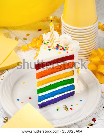 Slice of colourful birthday cake with lit birthday candle and yellow party decorations in the background.