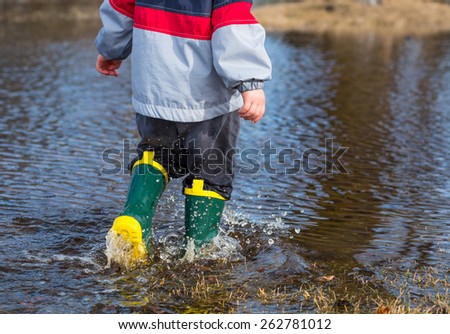 Very wet little boy strolling through puddles wearing rain boots.