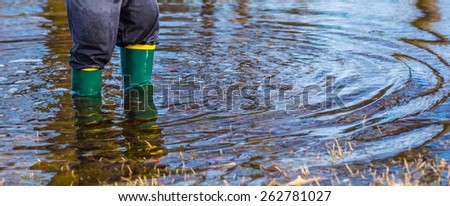 Very wet little boy standing in a puddle wearing rain boots.