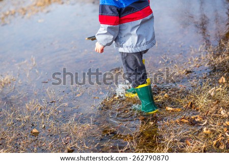 A little boy playing with a stick in puddles wearing rain boots.