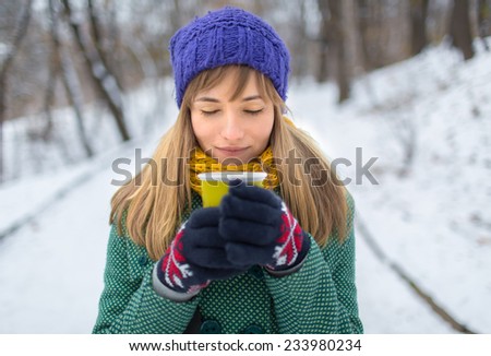 girl in green coat holding a cup of very hot coffee with steam with milk. Lonely woman stands on a snowy winter deserted street in the park.