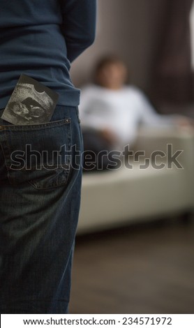 A man stands facing his pregnant partner. In his back pocket, her ultrasound peeks out. She is standing in the background holding her belly and out of focus.