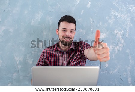 man with thumbs up - working at a laptop on the background of blue wall. Hispanic is playing on a laptop. Latino communicate in social networks