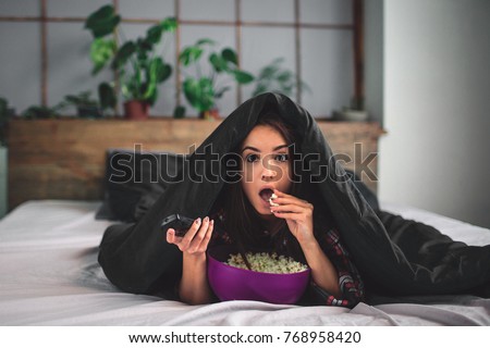 Young woman at home sofa couch in living room watching television scary horror movie or suspense thriller film or horrible news horrified holding remote controller in panic eating popcorn bowl