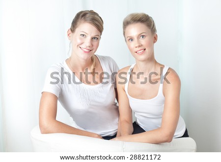 Portrait of two happy caucasian sisters sitting side-by-side shot against a cozy overall white home background