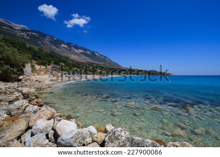 A beautiful beach stretches out from a group of rocks into clear blue water. In the peaceful village of Lourdas, on the island of Kefalonia, Greece.