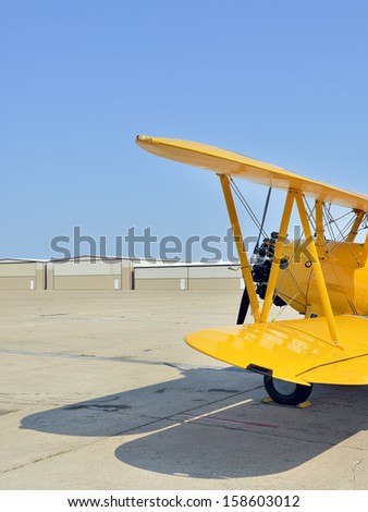 Old yellow biplane on empty runway with hangars behind