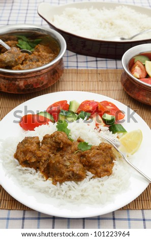 A plate of Madras butter beef curry with rice, salad and serving dishes in the background