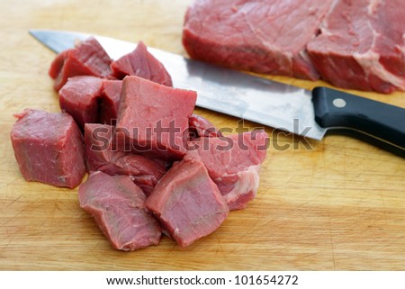 A chopping board with pieces of rump steak cut into cubes and another piece of steak in the background