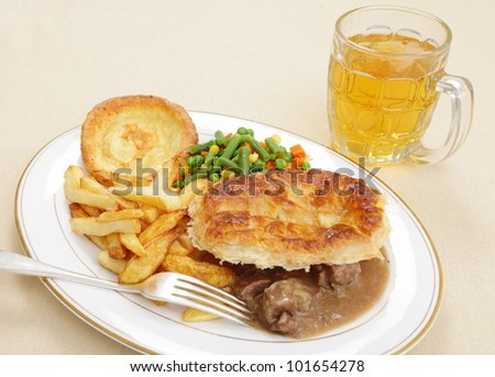 A meal of a homemade steak and kidney pie with french fried potato chips,yorkshire pudding and mixed vegetables served with a pint of lager beer