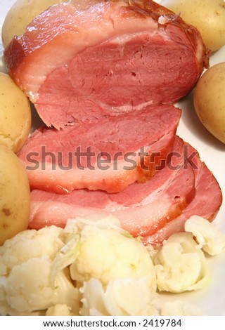 A serving dish with a baked gammon ham joint, sliced, boiled new potatoes in their jackets and boiled cauliflower