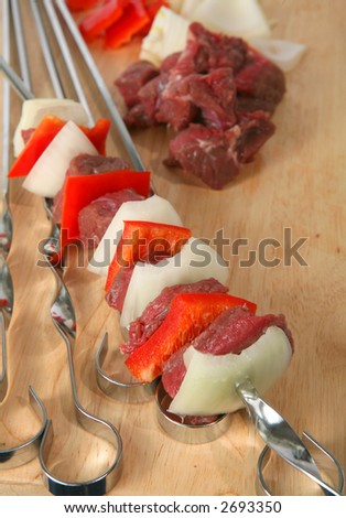 A kebab skewer with meat in the background, and other unused skewers beneath it.
