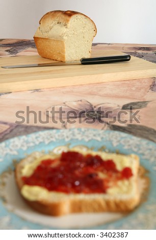 Breadboard with bread and carving knife. Out of focus slice of bread and jam in the foreground.