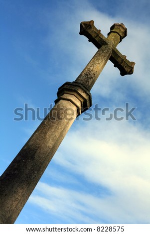 A huge carved stone cross against a blue and white sky. The cross is mottled with lichen and moss. Focus is in the upper part of the cross.