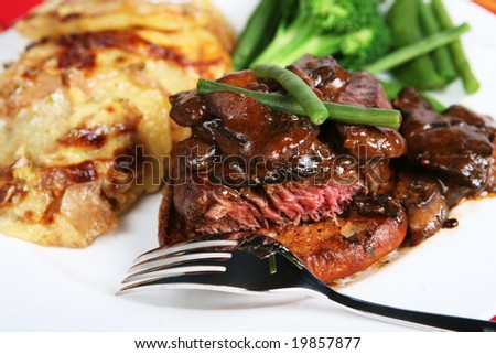 A meal of beef tournedos on a crouton, topped with mushrooms in a brown sauce and served with beans, peas and onion and potato gratin.