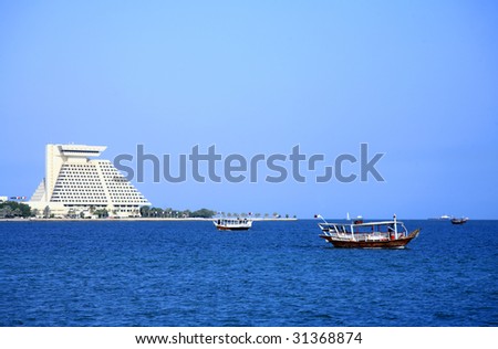 Three dhows ferrying sightseers ply the waters of Doha Bay, Qatar, Arabia, while the bulk of a landmark hotel looms behind them.