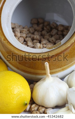 A rustic pottery storage jar with chickpeas (garbanzo beans) in it, lemon garlic and spilled chickpeas in the foreground. The three ingredients are used together in various recipies, such as hummus.