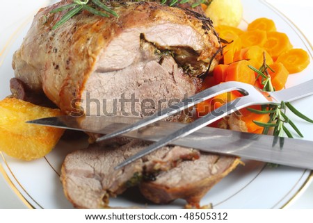 A serving plate with a joint of roasted boneless lamb roasted potatoes and boiled carrots, garnished with sprigs of rosemary with carving implements