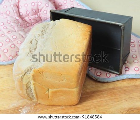 A loaf of English split-tin bread, fresh from the oven, on a wooden board with the baking tin and oven gloves.
