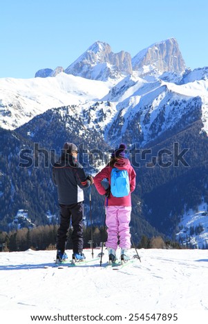 young girl in a bright track suit and a man on skis in the winter mountains in the background
