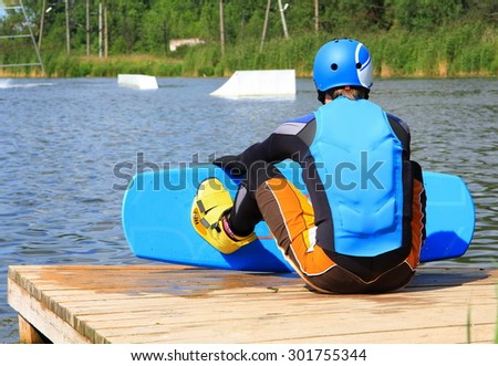 man -wake-boarder: black sports hydro-suit, blue life jacket, blue helmet on head, yellow shoes and blue board for wake-board sitting on a wooden quay on the shore of the lake in the background