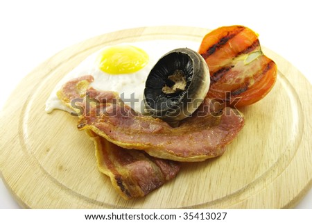 Slices of crispy pork bacon with half a grilled tomato a fried egg and a mushroom on a wooden round plate with a white background