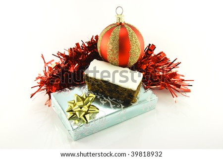 Slice of christmas cake with fruit filling and icing with a silver shiny gift, red bauble and red tinsel on a reflective white background