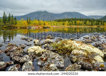 Rischorr mountain with peak hidden in clouds reflected in shallow Polygonal northern taiga forest lake with lichen-covered rocks in foreground