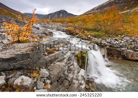 Small waterfall on Risjok freshwater stream with cloudy sky and tundra mountain slopes in background