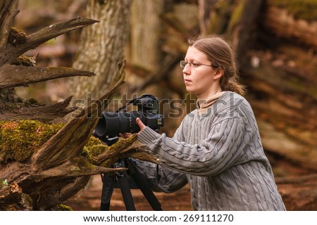 Woman takes a photograph of tiny plant between roots of a huge oak tree trunk using tripod and professional photo camera