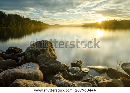 Long exposure shot of quiet sunset with moving clouds, forest, and still water in background, and with big rocks and pebbles on the foreground