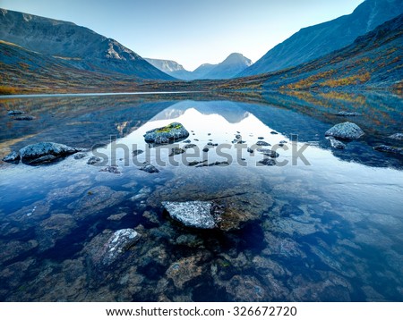 Landscape of lake Tahtarjavr with transparent water, rocky bottom and distant mountains reflected in still morning waters, Hibiny mountains above the Arctic circle, Russia