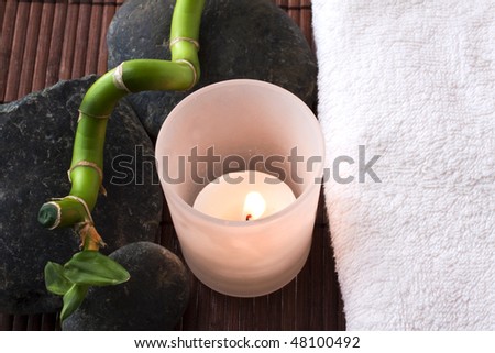 A white candle burns between white towel, bamboo branch and stones