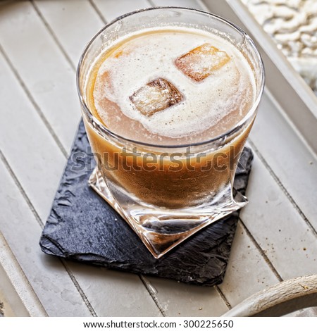 Drink in a glass over black stone coaster on a tray, with two ice cubes, square image