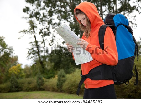 a beautiful caucasian young woman with map in her hands, rucksack on her back standing in a park outdoors in windy day on vacation