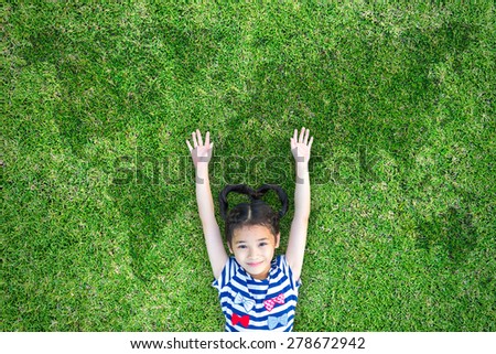 Happy healthy kid lying on green grass field raising two hands up with world map in the background : Little Asian child open arms with happiness in clean environment : Children in environment concept