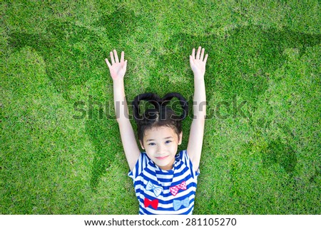 Happy Asian kid with heart-shaped hair raising hands up lying on green grass floor with world map background : World earth day with happy little kid : Save the planet and healthy environment concept