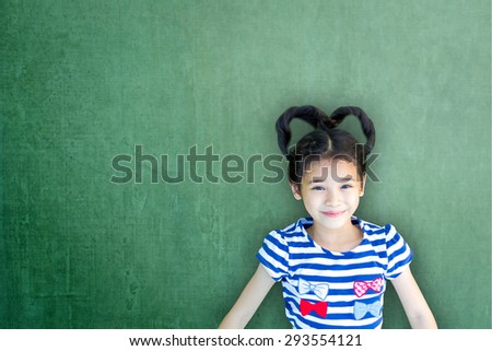 Happy Asian school child girl with heart shaped hair on green color chalkboard background : Smiling lovely female little kid with empty copy space on writable cement concrete backdrop for adding text