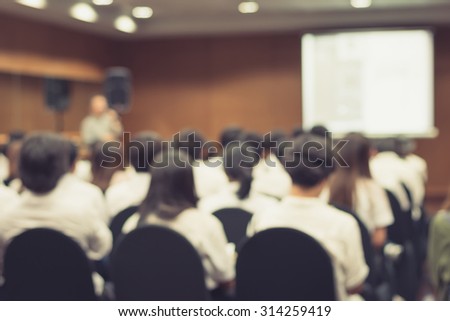 Vintage style blurred abstract background of university students sitting in a lecture room with teacher in front of the class with white projector slide screen: Blurry view from back of the classroom