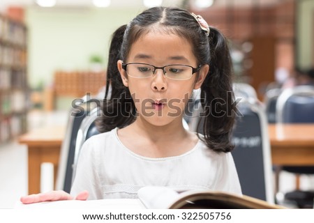 Little asian child girl wearing eyeglasses excited reading book in school library (Selective focus): Happy young female student kid looking at fairytale book with excitement in college study room