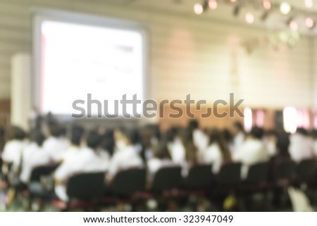 Blurred abstract background of business/ educational conference and seminar in auditorium hall with audiences/ students sitting in seat rows and presenters on stage with projector screen presentation