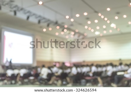 Blurred abstract background of business/ educational conference and seminar in auditorium hall with audiences/ students sitting in seat rows and presenters on stage with projector screen presentation