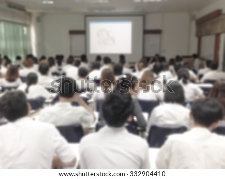 Blurred abstract background of college students sitting in seating rows attending school course: Blurry view from back of the classroom toward projector screen presentation in front of the class