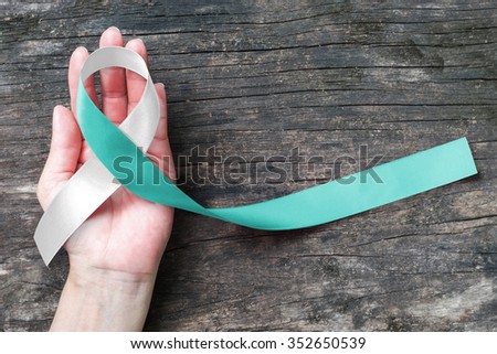 Teal & white ribbon on human hand on wood background to support persons with cervical cancer: Satin silk fabric symbolic concept for raising awareness/ campaign helping people living with illness