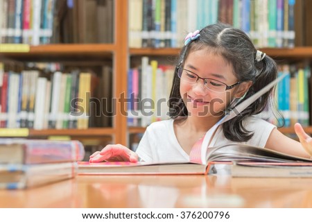 Happy little asian child girl w/ eyeglasses reading book school background: Lovely cute young female student kid opening flipping book in archive resource collection room: National library lover month