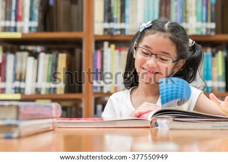 Happy little asian child girl w/ eyeglasses reading book school background: Lovely cute young student kid opening flipping book in archive resource collection room: National library lover month week